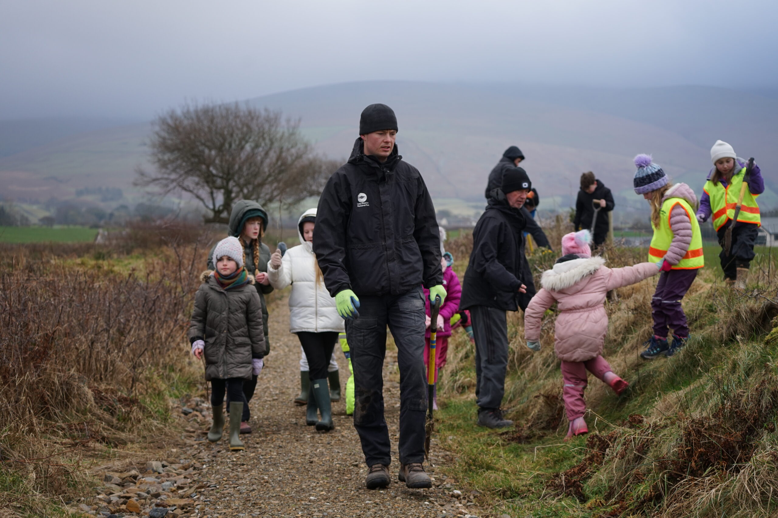 lake district national park authority ranger surrounded by children in outdoor clothing on a cloudy day in a field, planting young trees
