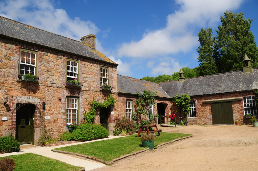 large rural country house made of red brick, slate roof and white cross sash windows. there is a main two storey house, with converted single storey barns all facing into a central yard with a loose stone driveway and well kept grass, with plants and flowers