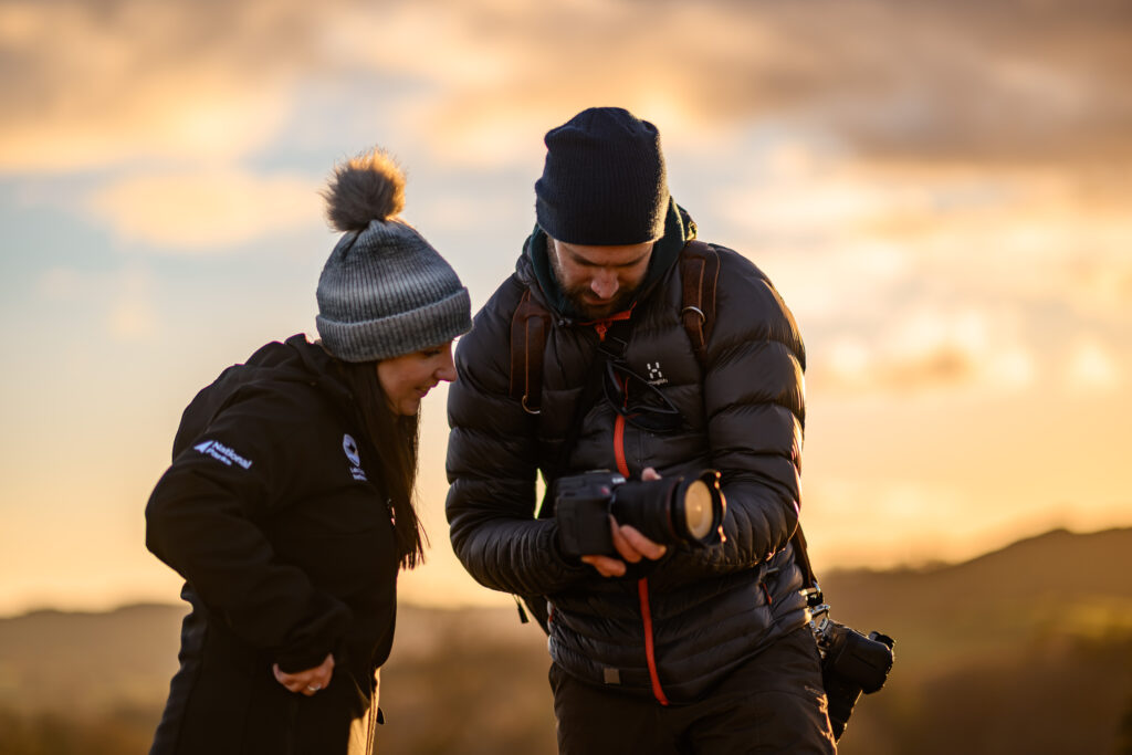 An adult woman and male in outdoor gear looking over a photography camera at sunset
