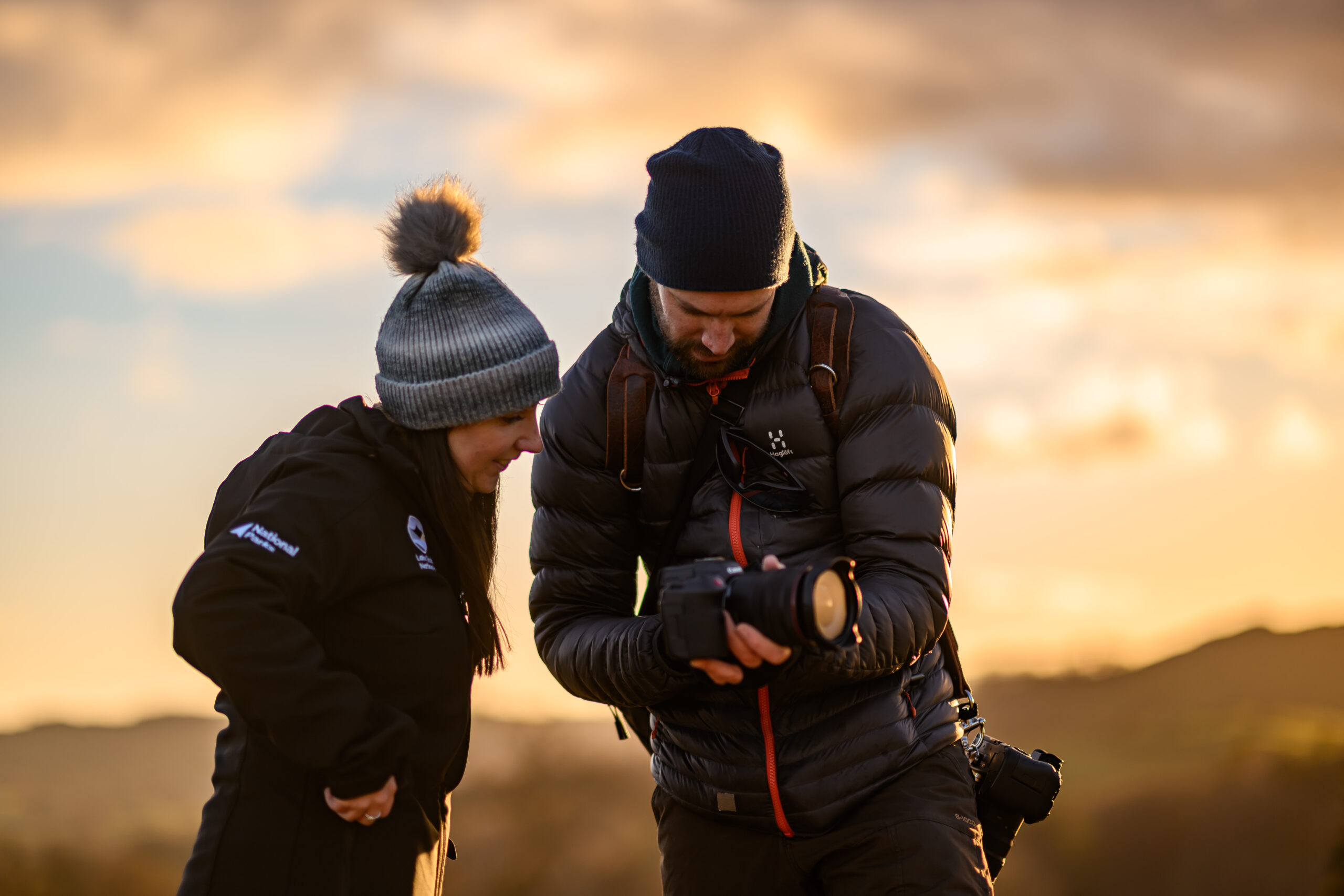 adult woman and male in outdoor gear at sunset looking over a photography camera
