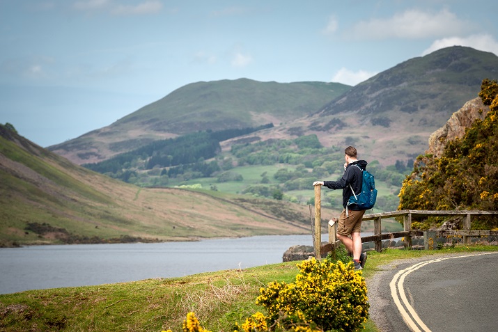 Walker standing on the side of a road, looking out over lake with mountains in the background.
