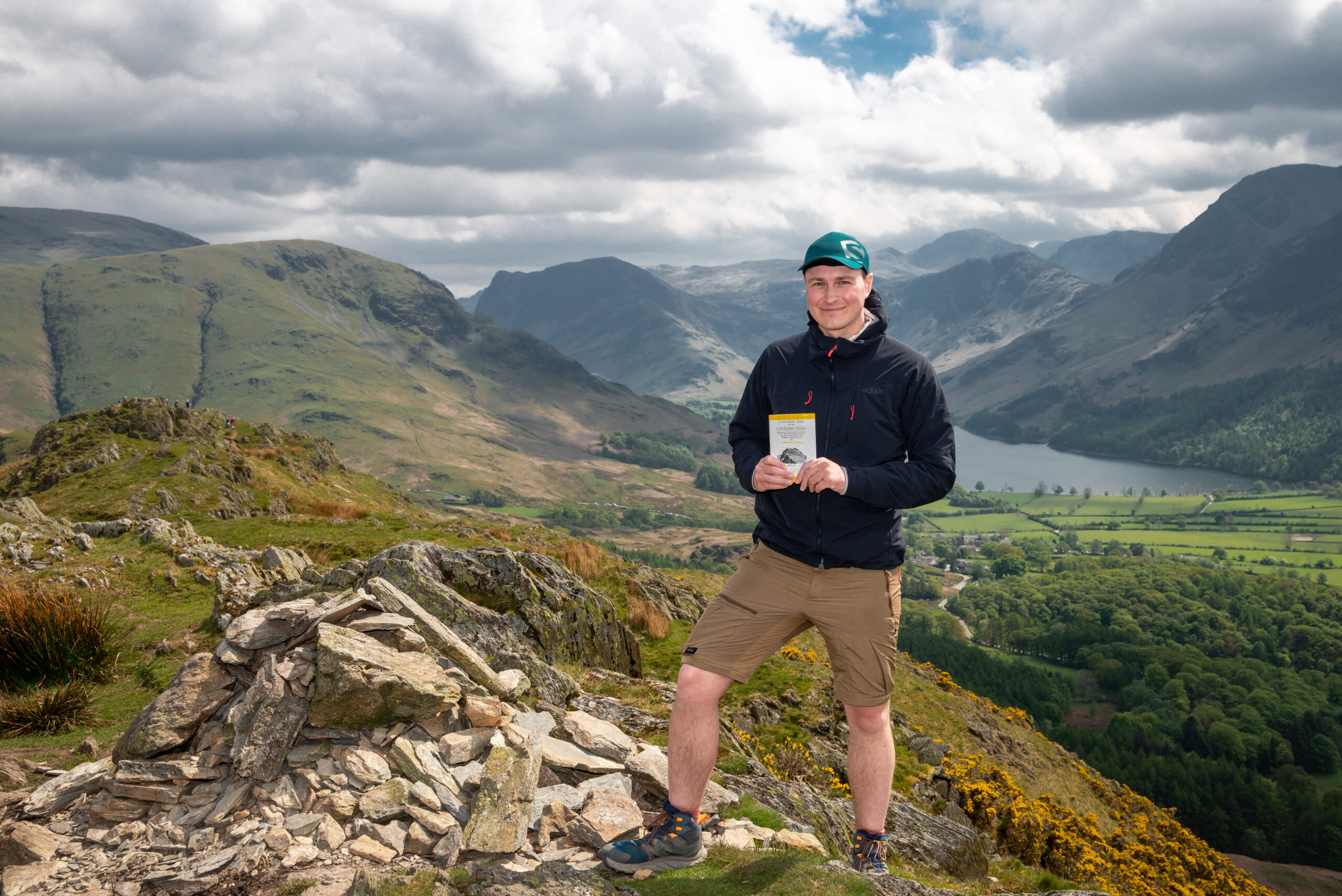Man holding a copy of Alfred Wainwright's book 'A Pictorial Guide to the Lakeland Fells' at the top of Rannerdale Knotts, with backdrop of the fells and large body of water.