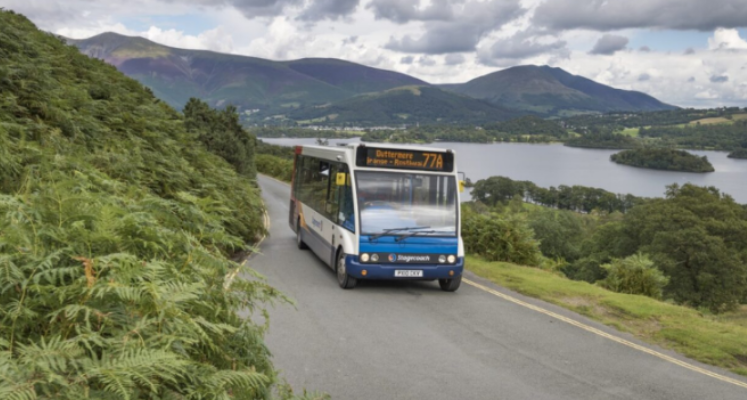 A bus driving through the Lake District with the lake and hills, trees and landscape in the background
