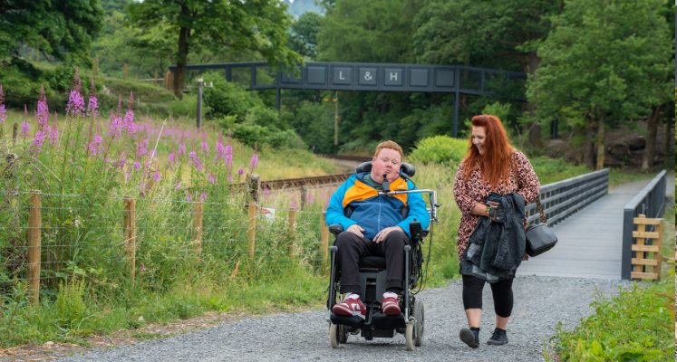 Young man in wheelchair with women walking along side him on a path with mountain backdrop