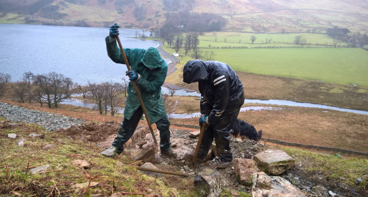 Two people in raicoats bent over looking at and working on a stone path. Behind them a body of water, and they are on the side of a fell