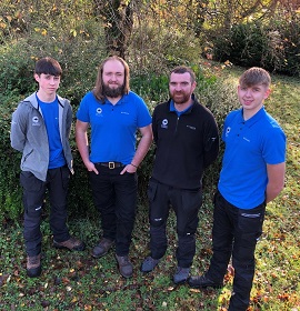 Four people in blue and black tops standing in a wooded area smiling at the camera