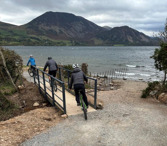People cycling over flat bridge with lake in background