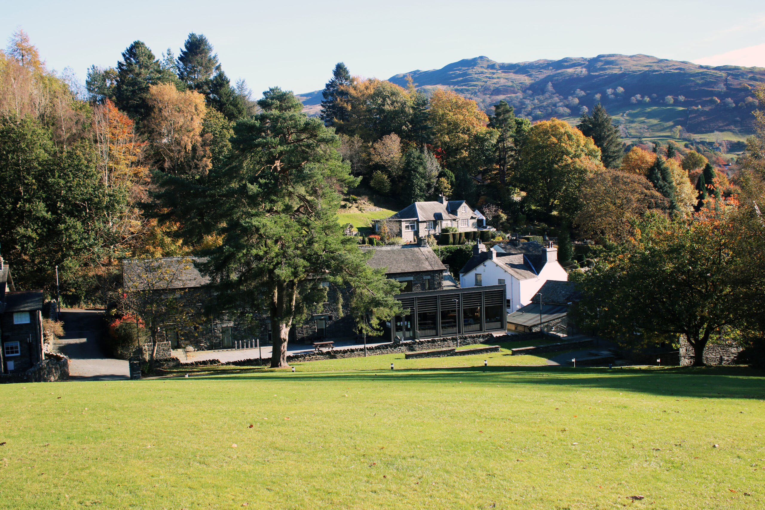 landscape view of large Ambleside University buildings nestled in woodland. Green grass field in foreground and fells in the backdrop