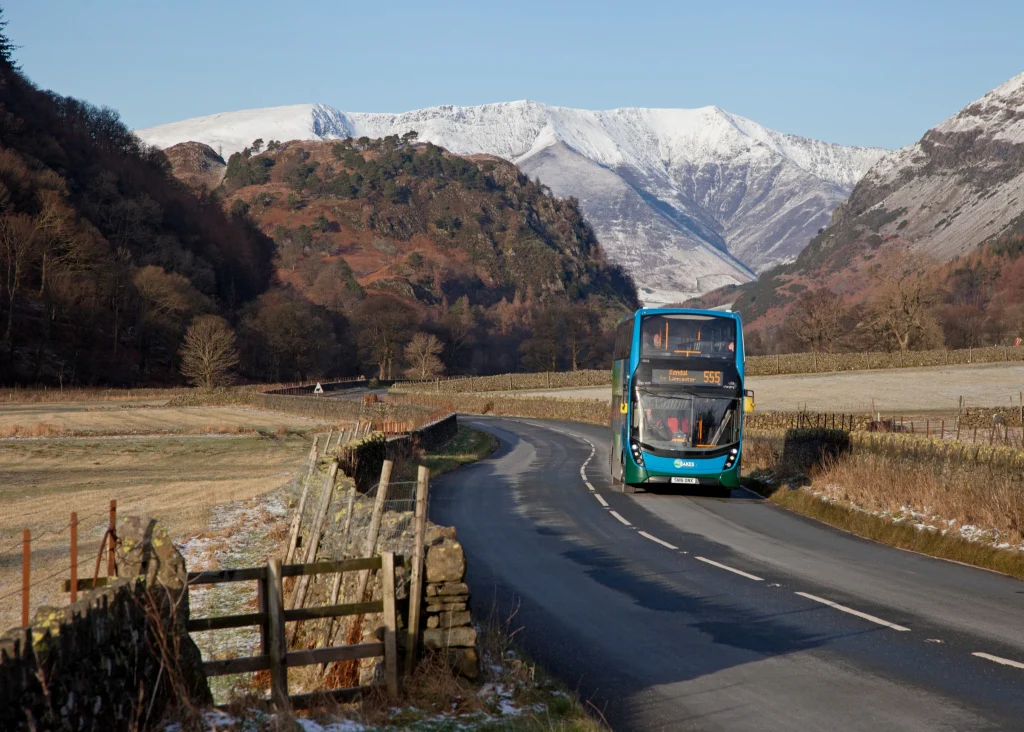 double decker bus on a road through the countryside with snow capped fells in the back drop