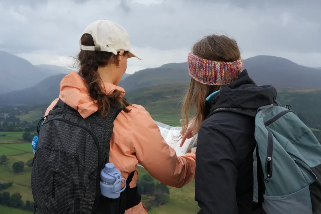 two young women facing away from camera looking out to misty fells in outdoor walking gear pointing at a map